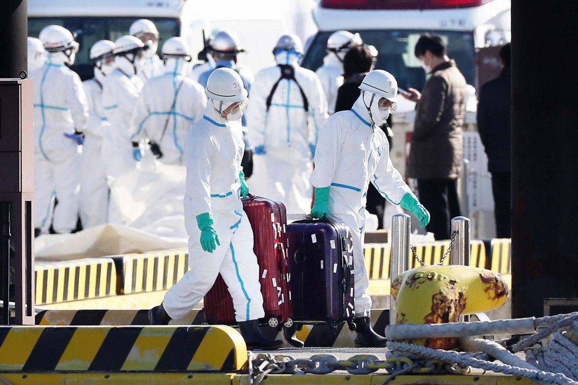 身著防護裝備的衛生官員從停泊在日本橫濱的鑽石公主號遊輪上帶出行李箱，大眾普遍認為這些行李箱的主人，就是驗出冠狀病毒陽性後從船上撤離送醫的乘客。PHOTOGRAPH BY ASAHI SHIMBUN, GETTY IMAGES