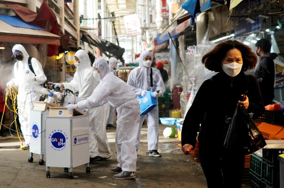 2020年2月26日，消毒公司的員工在幫南韓首爾一處傳統市場消毒，旁邊一名女性戴著口罩，以免感染新型冠狀病毒。PHOTOGRAPH BY KIM HONG-JI, REUTERS