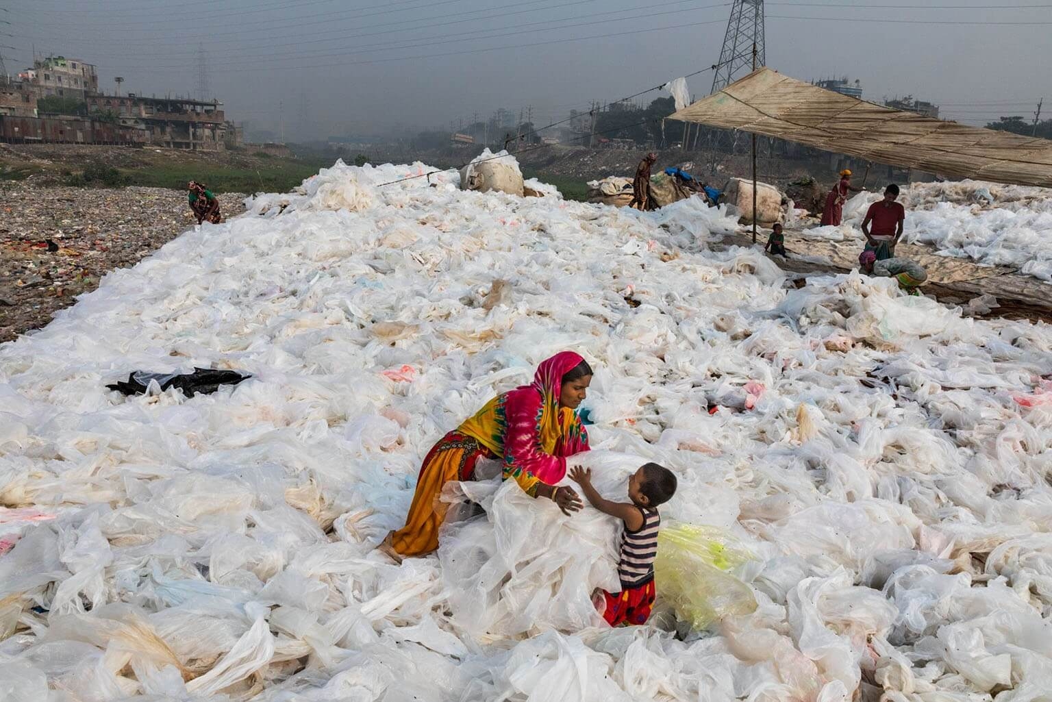 一張張透明塑膠垃圾在孟加拉達卡的布里甘加河中洗淨後，諾亞涵把它們攤開來曬乾，定時翻動，一邊照顧兒子莫莫。這些塑膠垃圾最後會賣給回收業者。全球生產的塑膠只有不到五分之一被回收，在美國的比例則不到10%。攝影：藍迪．奧森 RANDY OLSON