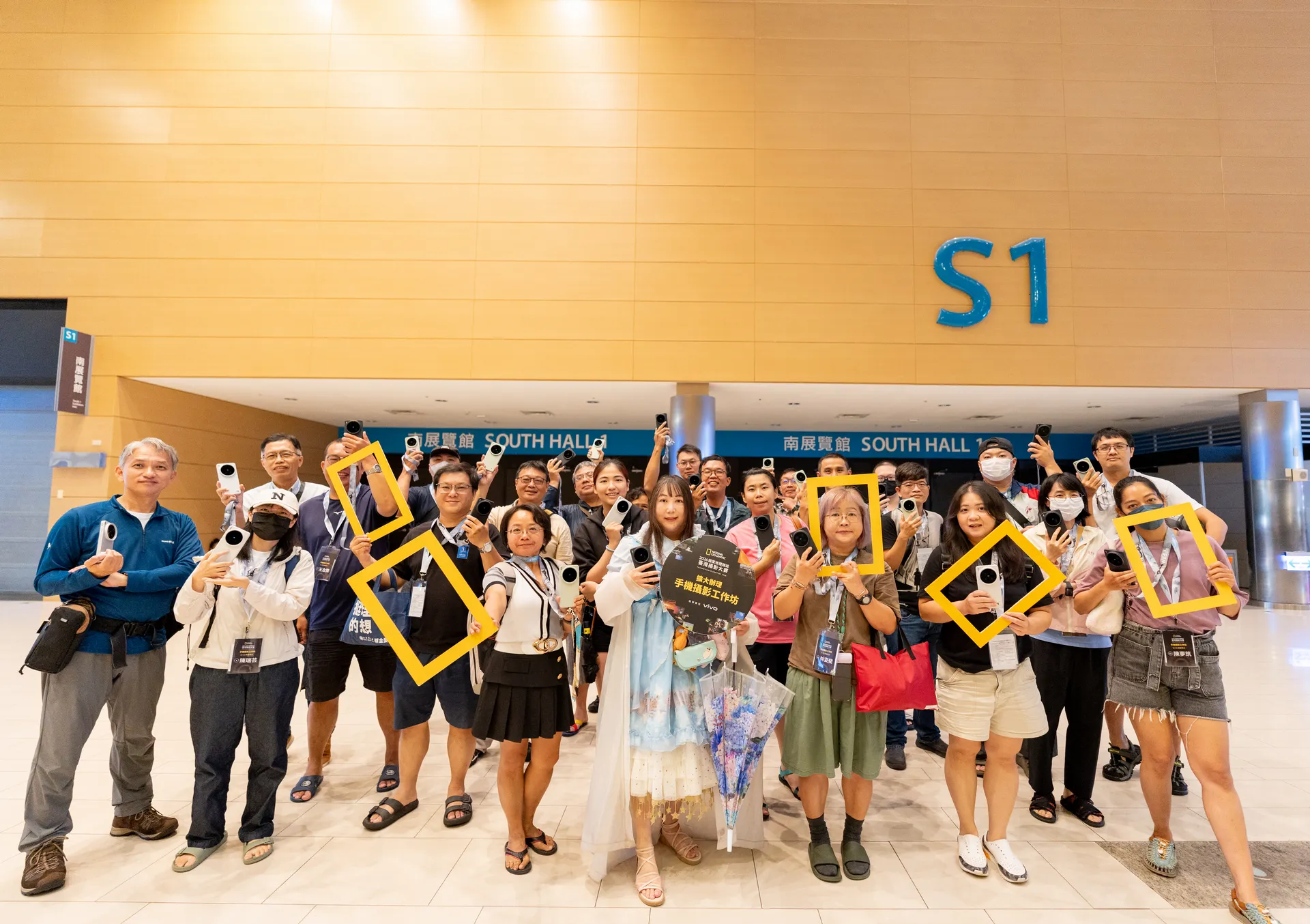 8月23日的高雄場學員因天候驟雨，改於高雄展覽館借用空間進行參觀，學員手持手機與《國家地理》黃框於入口前合影留念